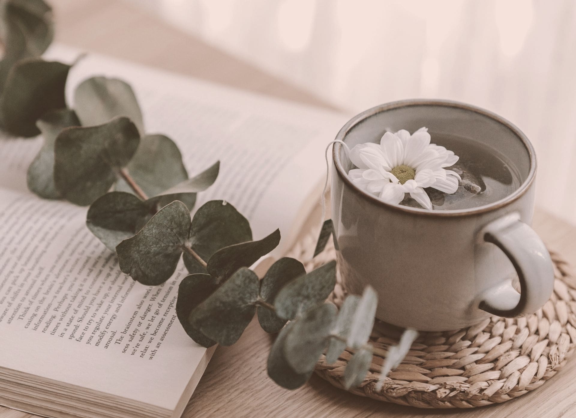 Table avec une tasse de thé avec fleur blanche sur un set de table en rotin, un livre ouvert et une branche dans un esprit Hygge.